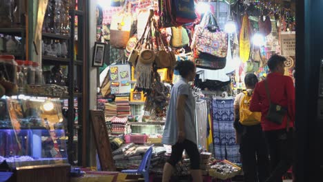 Plano-Exterior-Medio-De-Personas-Caminando-Lentamente-Por-Las-Tiendas-Del-Mercado-Nocturno-Con-Una-Dama-Saliendo-Del-Puesto-Por-La-Noche