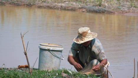 Fisherman-in-Straw-Hat-Crouching-Down-Putting-The-Last-of-His-Catch-In-His-Bucket-Then-Standing-To-Shake-Out-The-Last-of-it-From-His-Net