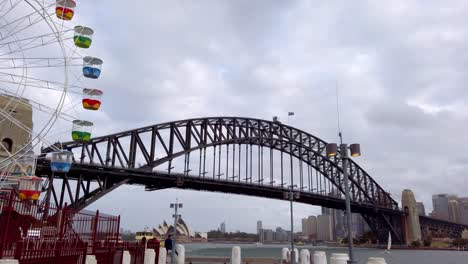 Parque-De-Diversiones-Luna-Con-Carrusel-Girando-Mientras-La-Vista-Cambia-Al-Puente-Del-Puerto-Y-Al-Horizonte-En-Un-Día-Nublado,-Tiro-Panorámico-A-La-Derecha