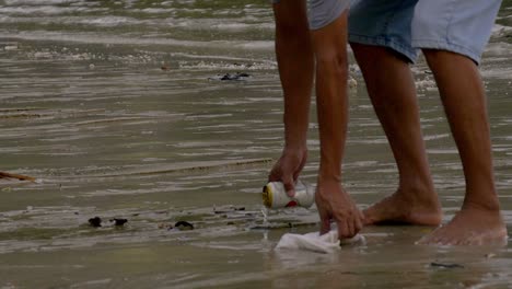 A-man-in-his-shorts-picking-up-garbage-washed-on-the-beach---Close-up