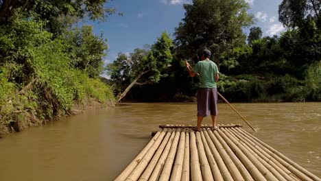Thai-man-steers-a-bamboo-raft-down-a-brown-flowing-river,-in-Northern-Thailand