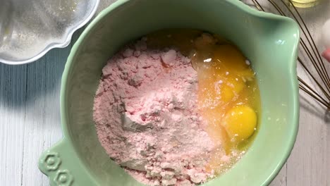 closeup-of-child's-hands-preparing-a-pink-cake-batter-by-adding-ingredients-and-cracking-eggs-in-top-view