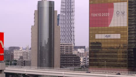 Publicidad-En-Vallas-De-Los-Juegos-Olímpicos-De-Tokio-2020-En-Tokio-Con-Tráfico-De-Automóviles-Y-La-Base-Del-árbol-Del-Cielo-Como-Telón-De-Fondo