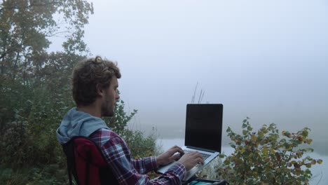 Ruhige-Aufnahme,-Ein-Mann,-Der-Einen-Laptop-Benutzt,-Neblige-Bäume-Und-Ein-See-Im-Hintergrund