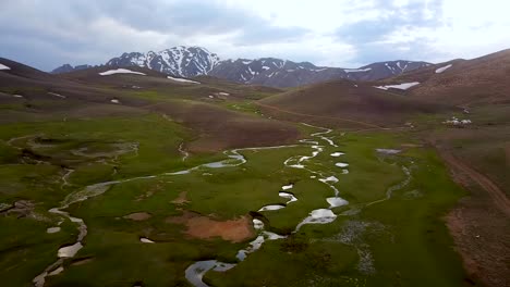 Grünes-Hochland-Aus-Der-Luft-Mit-Schneebedeckter-Berglandschaft-Und-Wunderschönem-Bach-Spiegelt-Den-Bewölkten-Himmel-An-Frühlingstagen-In-Kerman,-Iran-Wider
