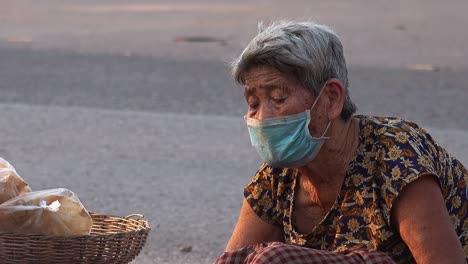 Close-Shot-a-Lady-Sitting-at-The-Side-of-a-Road-With-a-Face-Mask-Looking-Down-Doing-Something-While-Motorcycles-go-Past-in-the-Background