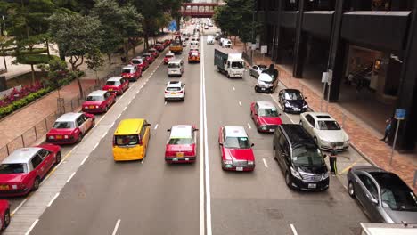 Tráfico-Que-Pasa-En-El-Centro-De-Hong-Kong-Con-Típicos-Taxis-Rojos-Estacionados-Al-Lado-De-La-Carretera,-Vista-Aérea-De-ángulo-Bajo
