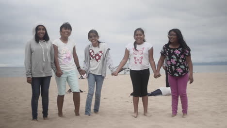 Group-of-Friends,-Elementary-Students,-Girls-Smile-to-Camera-and-Hold-Hands-at-the-Beach,-Slow-Motion