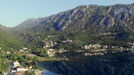 Eine-Riesige-Gebirgskette-Rund-Um-Die-Bucht-Von-Kotor,-Montenegro,-Die-über-Einer-Kleinen-Küstenstadt-An-Ihren-Ufern-Aufragt,-Die-Berge-Spiegeln-Sich-Im-Klaren-Wasser-Darunter,-Beleuchtet-Von-Goldenem-Sonnenlicht,-Luftaufnahme