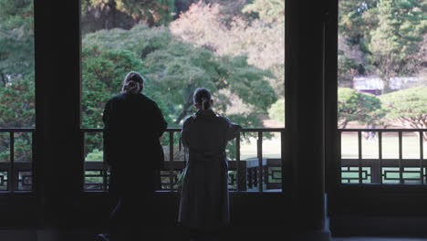 Dos-Mujeres-Japonesas-Admirando-La-Vista-En-Un-Jardín-Japonés-Desde-Un-Pabellón-En-Tokio,-Japón---Plano-Medio
