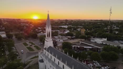 Orbital-parallax-of-neo-gothic-church-and-tower-leading-to-boulevard-main-street-in-Santa-Elisa-countryside-town-at-sunset,-Entre-Rios,-Argentina