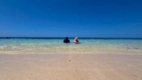 Couple-Sitting-Together-In-Ocean-As-Waves-Gently-Go-Past-Them-Near-Beach