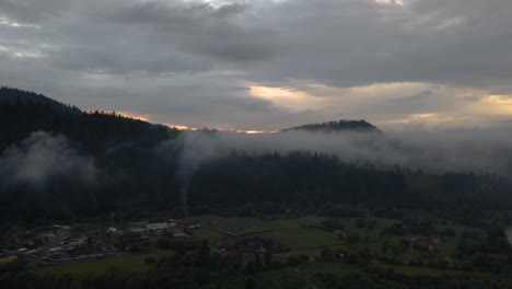 Smoke-rising-from-chimneys-in-a-small-village-surrounded-by-hills-covered-by-forests,-flying-up-to-reveal-beautiful-golden-sunrise