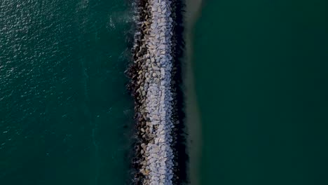 Birdseye-view-of-rock-wall-ocean-jetty-creating-a-sand-bank,-sunny-day