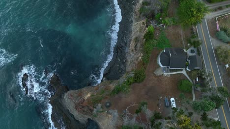 Top-down-aerial-view-of-a-steep-cliffside-lining-California's-scenic-highway-with-a-house-overlooking-the-ocean