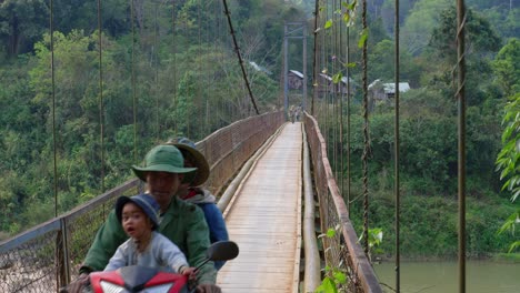 Familia-Asiática-Con-Niño-En-Ciclomotor-Cruzando-Madera-Y-Puente-De-Cuerda-Sobre-El-Río,-Vietnam