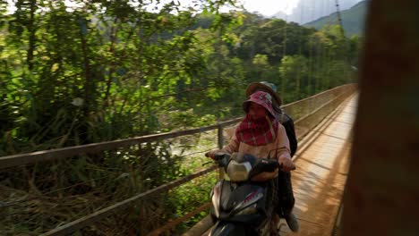 Asian-couple-riding-moped-across-a-river-bridge-and-up-a-hill-on-dirt-road,-Vietnam