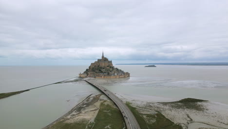 People-walking-and-bus-driving-along-bridge-of-Mont-Saint-Michel-during-low-tide-on-cloudy-day,-Normandy-in-France