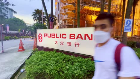 Nahaufnahme-Eines-Banners-Einer-öffentlichen-Bank-Vor-Einem-Hohen-Gebäude-In-Kuala-Lumpur,-Malaysia,-Mit-Vorbeikommenden-Einheimischen-Am-Abend