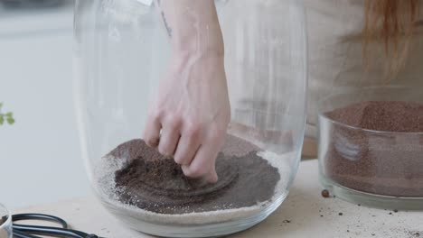 A-young-woman-creates-a-small-ecosystem-in-a-glass-terrarium---preparing-the-soil-layer---nature-preservation-concept---medium-shot