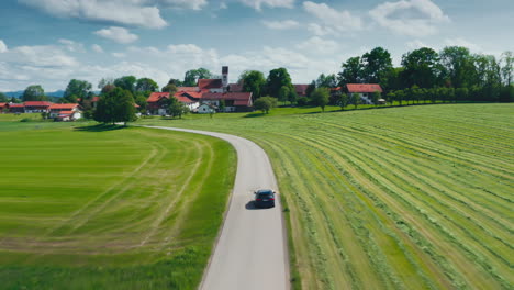 High-drone-shot-of-a-blue-BMW-car-driving-down-a-scenic-road-towards-a-small-viallage-surrounded-by-lush-green-meadows-on-a-sunny-day