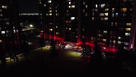 Camiones-De-Bomberos-Fuera-De-Un-Edificio-De-La-Ciudad,-Con-Luces-De-Sirena-Encendidas-Y-Mucha-Gente-Alrededor,-Por-La-Noche