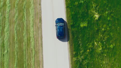 Topshot-of-a-blue-BMW-car-driving-through-countryside,-surrounded-by-green-meadows-in-breathtaking-drone-footage