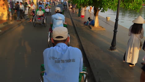 Traditionelle-Vietnamesische-Fahrradrikschas,-Die-Touristen-In-Der-Engen-Straße-Der-Altstadt-Von-Hoi-An-Befördern,-Während-Andere-Menschen-Den-Sonnenuntergang-In-Der-Nähe-Des-Flussufers-In-Vietnam-Besichtigen-Und-Genießen