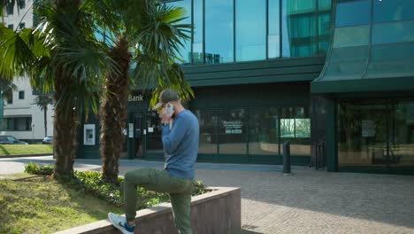 Male-Wearing-Baseball-Cap-And-Sunglasses-Pacing-Around-Taking-Phone-Call-Outside-Glass-Office-Building