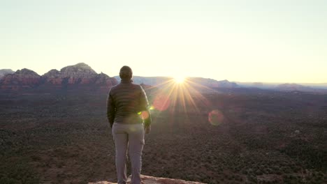 Hiker-stands-on-cliff-at-sunrise-watches-sun-crest-over-mountain-tops-in-Sedona