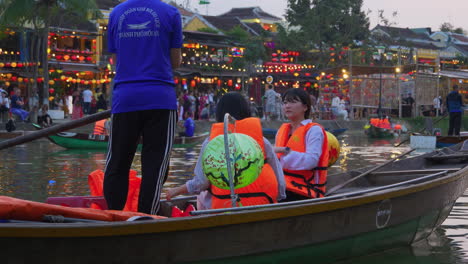 Touristen-Unternehmen-Abends-Eine-Laternenbootfahrt-Auf-Dem-Hoai-Fluss-In-Einem-Traditionellen-Sampan-Boot-In-Der-Antiken-Stadt-Hoi-An-Mit-Blick-Auf-Die-Stadt-Im-Hintergrund,-Vietnam