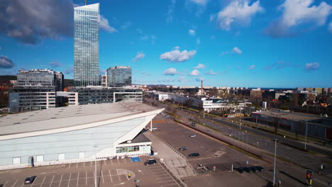 Aerial-Flying-Over-Car-Park-With-Large-Building-With-Sloped-Roof-And-Tall-Office-Building-In-Background-In-Przymorze