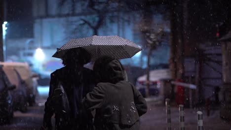 In-Schwarz-Gekleidete-Freunde-Teilen-Sich-Einen-Regenschirm-Und-Gehen-Weg,-Während-Nachts-Schnee-Fällt