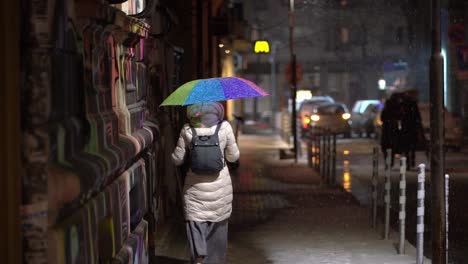Woman-in-white-coat-holds-rainbow-umbrella,-walks-under-street-light-at-night