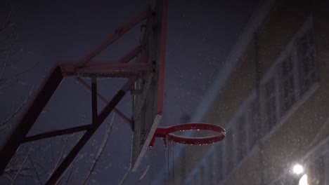 Snow-falls-around-basketball-backboard-and-rim-with-no-net-at-night