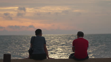 Zwei-Männer-Mittleren-Alters-Sitzen-Am-Meer-Und-Genießen-Den-Farbenfrohen,-Leuchtend-Rosafarbenen-Sonnenuntergang-über-Dem-Endlosen-Meer-In-Kota-Kinabalu-–-Rückansicht