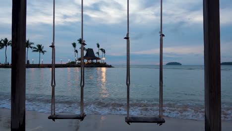 Wooden-swing-on-a-tropical-remote-dream-honeymoon-beach-at-the-seaside-with-small-islands-in-background-at-sunset,-Dramatic-courful-sky
