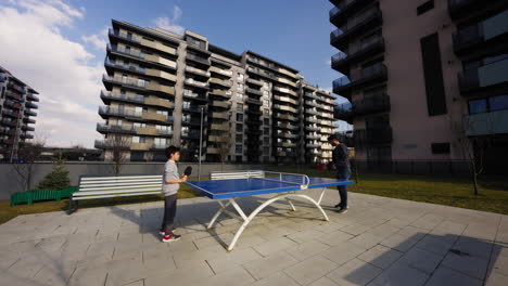 Two-people-playing-table-tennis-outside,-on-a-luxury-residential-apartment-complex-park,-on-a-sunny-day
