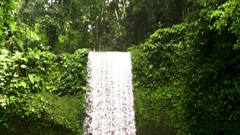 Downward-pan-over-stunning-jungle-Tibumana-waterfall,-people-swim-in-pool