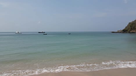 Low-aerial-leaves-sand-beach-to-sea-and-fishing-boats-in-bay,-Thailand