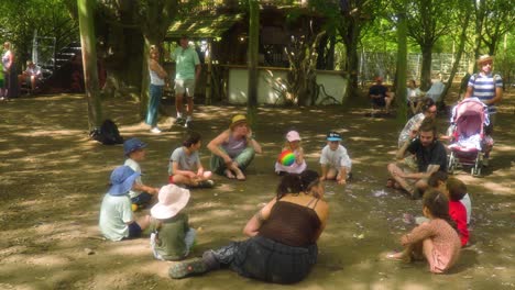 group-of-kids-with-adults-sitting-in-a-circle-playing-games-getting-to-know-each-other-in-a-family-area-on-a-summer-camp-festival-in-the-woods-with-parents-standing-around-enjoying-music