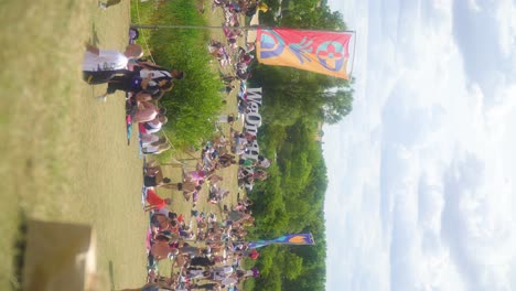 overview-of-the-summer-music-festival-We-Out-Here-people-relaxing-outdoor-in-nature-sitting-down-walking-around-in-the-field-beautiful-trees-in-the-background-and-colourful-artistic-flags-vertical
