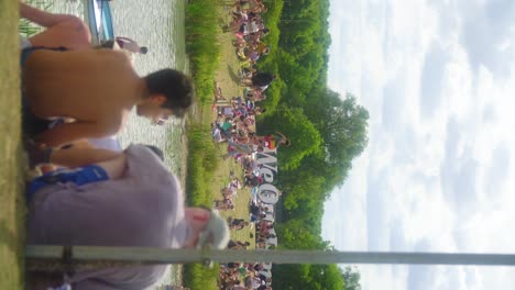 crowded-summer-music-festival-people-relaxing-by-the-lake-through-the-sunny-day-riding-on-a-boat-swimming-walking-around-to-get-food-dressed-in-swim-suits-nature-scenery-with-big-trees-vertical-video