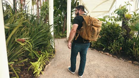 Young-male-traveler-with-backpack-walks-through-a-tropical-greenhouse-in-slow-motion