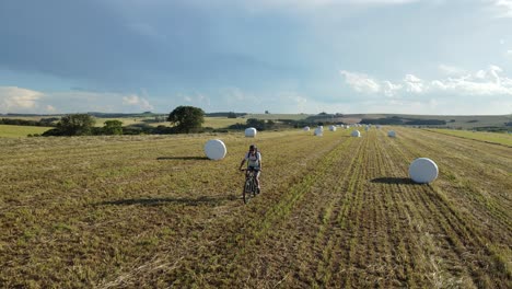 Cyclist-passing-between-silage-of-rolled-hay-wrapped-in-a-harvested-field,-drone-view