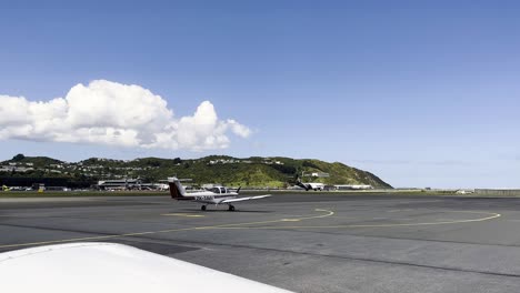 Single-engine-Aircraft-Parked-At-The-Apron-Of-The-Airport-With-Air-New-Zealand-PlaneTaking-Off-In-The-Background