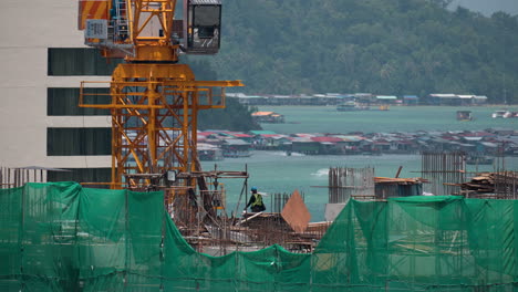 Arbeiter-Arbeiten-Auf-Der-Baustelle-Eines-Mehrfamilienhauses-Mit-Kran-Oben,-Wasserdörfer-Der-Stadt-Kota-Kinabalu-Bei-Der-Insel-Gaya-Im-Hintergrund---Nahaufnahme-In-Zeitlupe