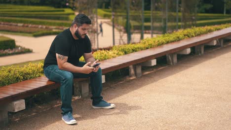 Young-man-pinking-up-and-texting-on-his-cell-phone-while-seated-on-a-long-bench-at-the-park-outdoors-in-slow-motion