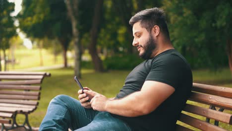 Young-man-using-his-mobile-phone-while-seated-on-a-park-bench-smiling-on-sunny-day