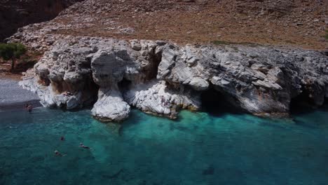 Menschen-Schwimmen-Im-Türkisfarbenen-Wasser-Und-Erkunden-Höhlen-In-Felsen-Am-Marmara-Strand,-Kreta,-Griechenland